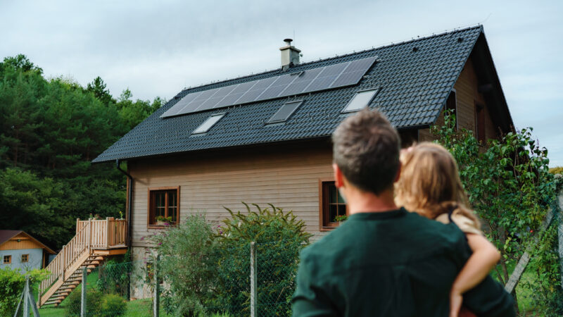 Rear view of dad holding her little girl in arms and looking at their house with solar panels.Alternative energy, saving resources and sustainable lifestyle concept.